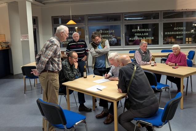 Skatturnier 2025 Punktezählung Die Teilnehmenden vom Skatturnier versammeln sich um einen Tisch, um der finalen Punktezählung zu folgen.